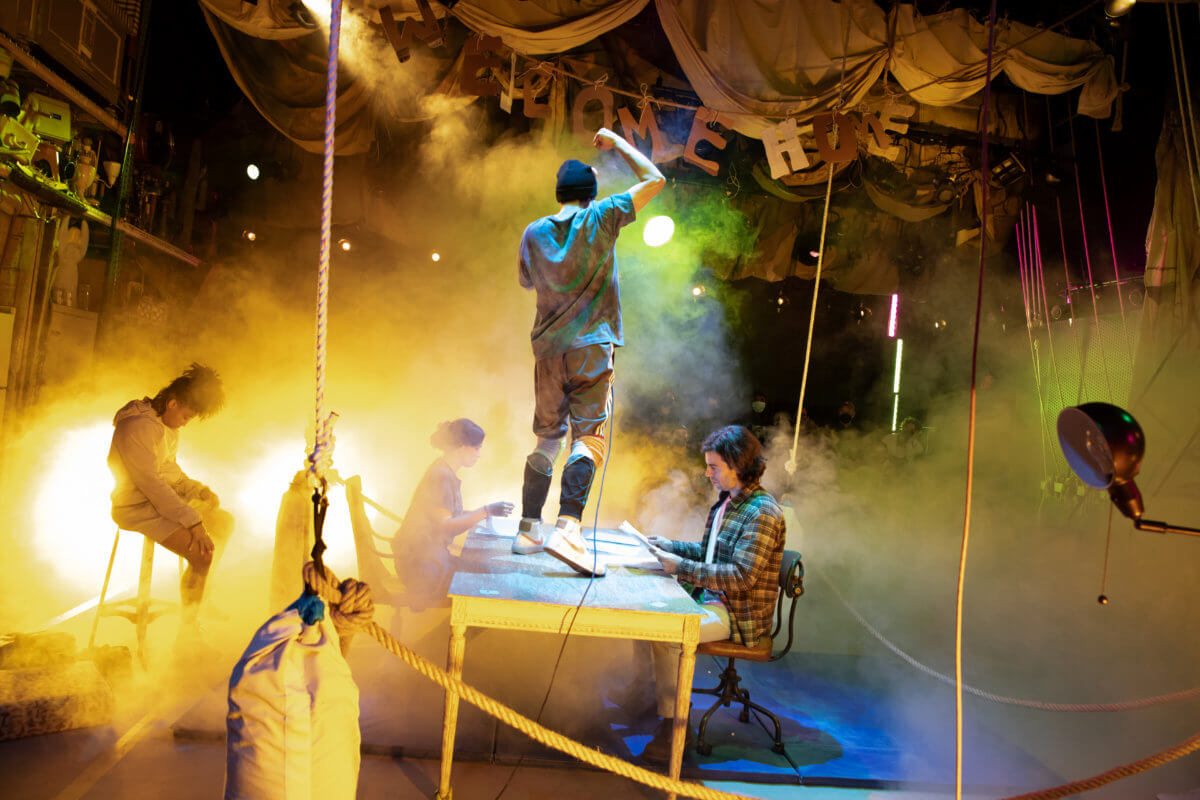 Four actors perform on a stage filled with fog and theatrical lighting. One sits off to the left on a stool, their head down. One stands on a table at center, facing away and holding a microphone. The other two sit at the table, opposite each other.